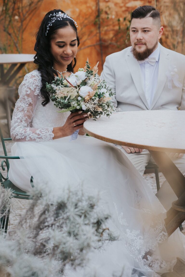 Frisch verheiratetes Paar in weißen Outfits sitzt an einem Tisch mit herbstlichen Bäumen im Hintergrund.