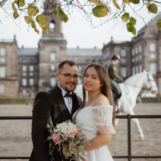 Heiraten in Dänemark: Ein Brautpaar posiert vor Christiansborg Slot mit einem Reiter auf einem weißen Pferd im Hintergrund.