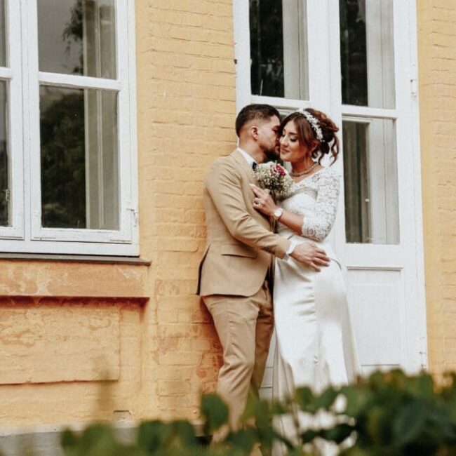 Hochzeitspaar posiert innig vor einem historischen gelben Gebäude mit weißen Fenstern.