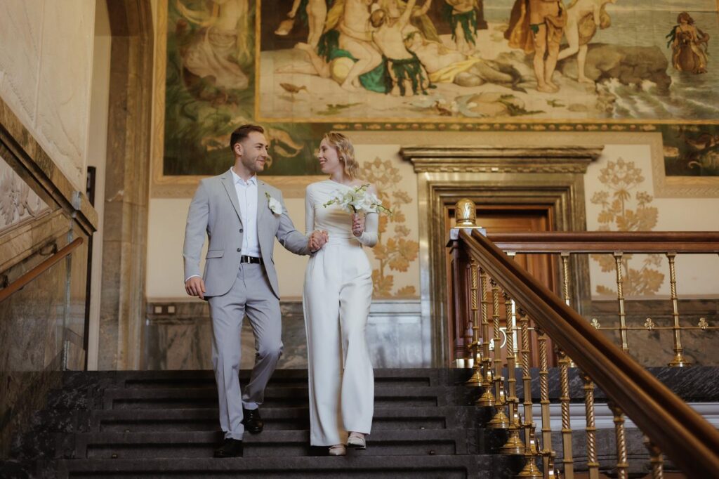 Heiraten in Dänemark: Ein glückliches Brautpaar geht die Treppe in einem historischen Gebäude mit kunstvollen Wandgemälden hinunter.