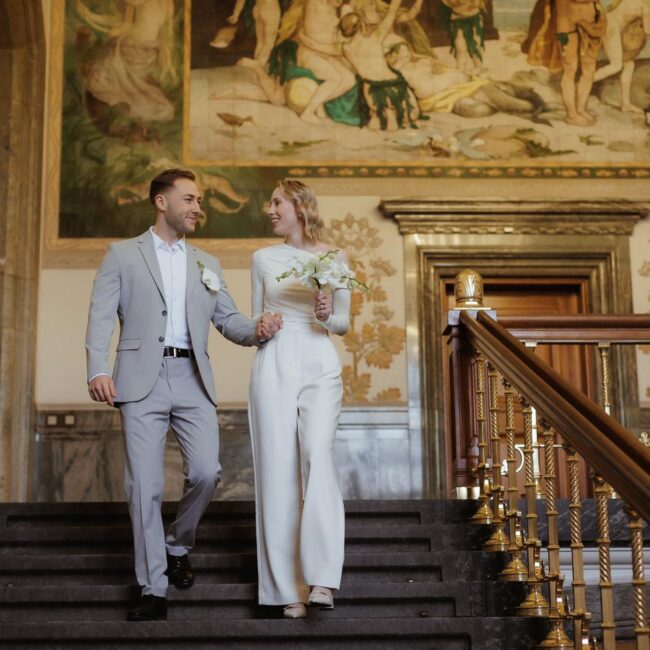 Heiraten in Dänemark: Ein glückliches Brautpaar geht die Treppe in einem historischen Gebäude mit kunstvollen Wandgemälden hinunter.