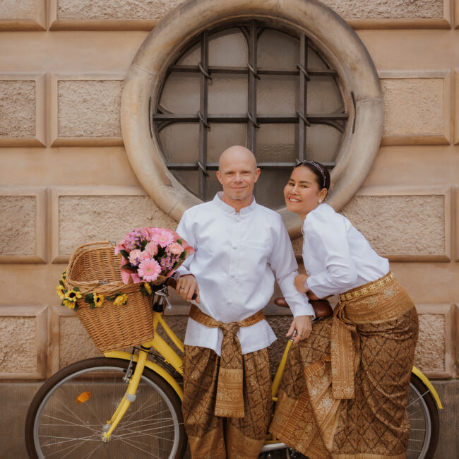 Heiraten in Dänemark: Ein Paar in traditioneller Kleidung posiert fröhlich mit einem gelben Fahrrad und Blumen vor historischer Kulisse.