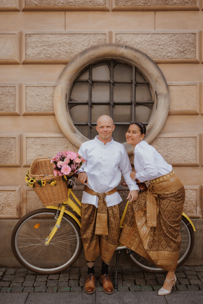 Heiraten in Dänemark: Ein Paar in traditioneller Kleidung posiert fröhlich mit einem gelben Fahrrad und Blumen vor historischer Kulisse.
