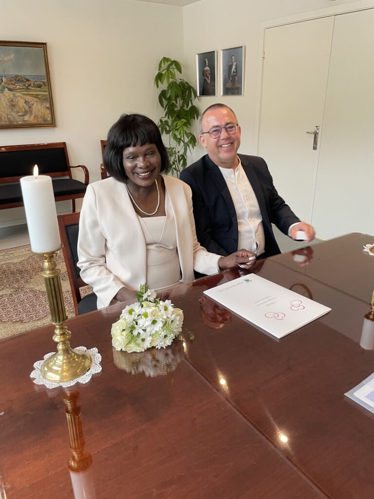 Ein glückliches Paar nach der standesamtlichen Hochzeit in Dänemark, sitzend an einem Tisch mit Blumenstrauß und Traudokumenten.